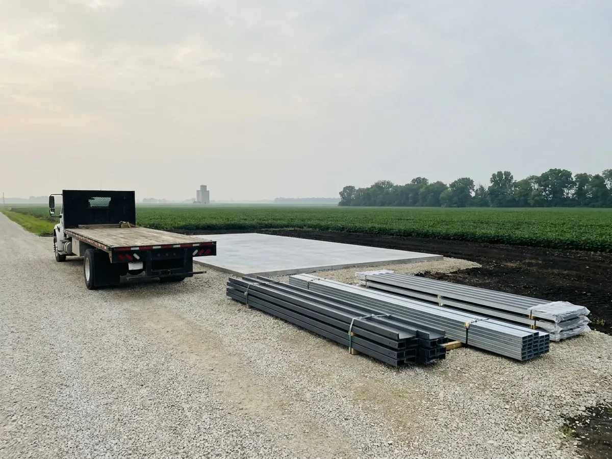 A flatbed truck delivering banded steel frame members and stacked metal roof panels to a building site beside a finished concrete slab.