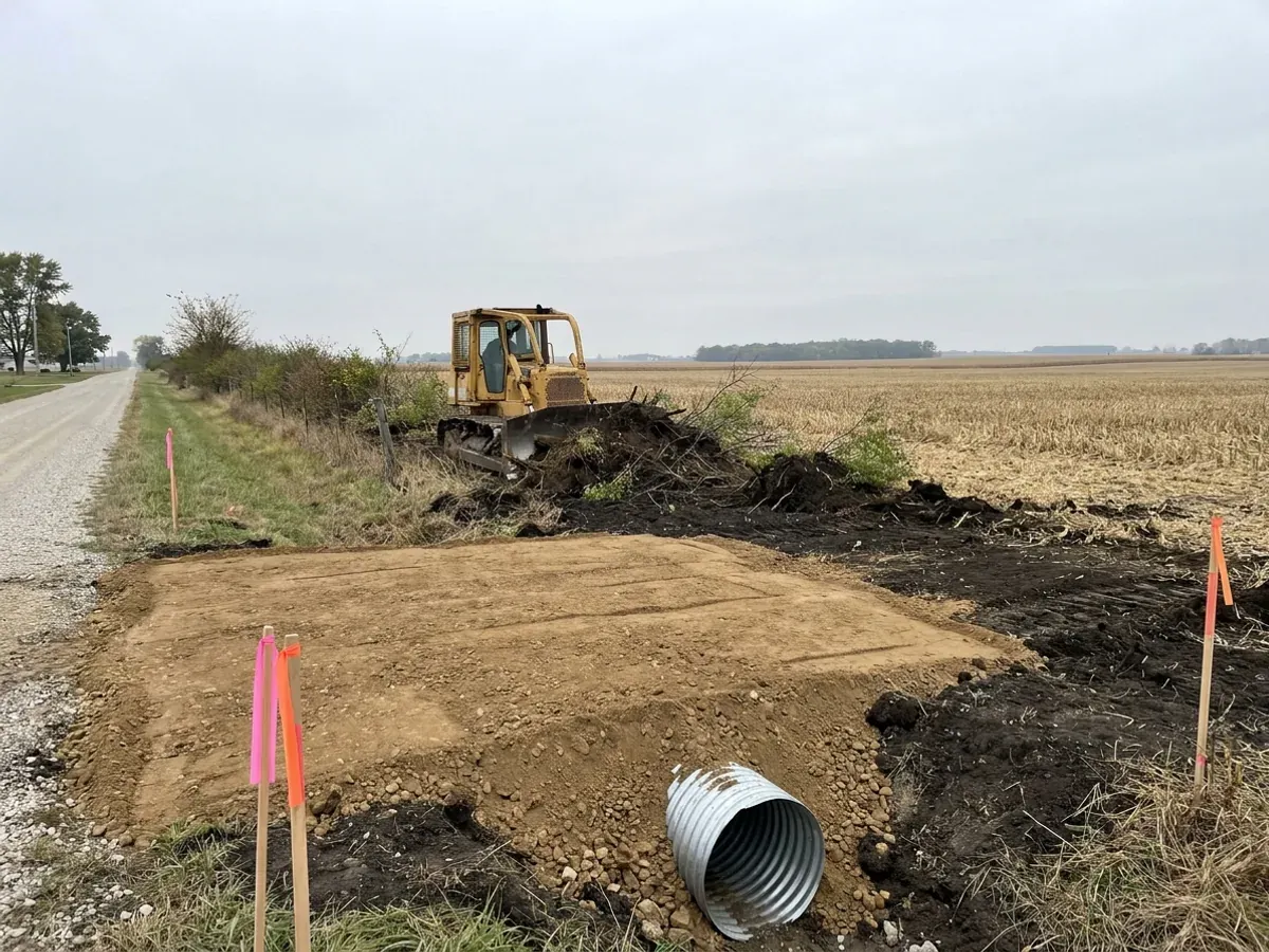 A dozer clearing an old fence-row beside a compacted crushed-stone building pad, with stripped black topsoil stockpiled and a culvert set at the driveway entrance.